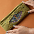 Load image into Gallery viewer, Green clutch with embroidered moth design held by hands on an orange background