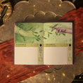 Load image into Gallery viewer, An open weekly planner spread with illustrated hands reaching toward olive branches, styled on rust-red fabric over an ornate metallic tray with warm string lights.
