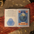 Load image into Gallery viewer, An open book spread showing printed pages titled 'The Forces at Play' and 'The Beaver Moon,' placed on a warm rust-red fabric over an ornate metallic tray with string lights.

