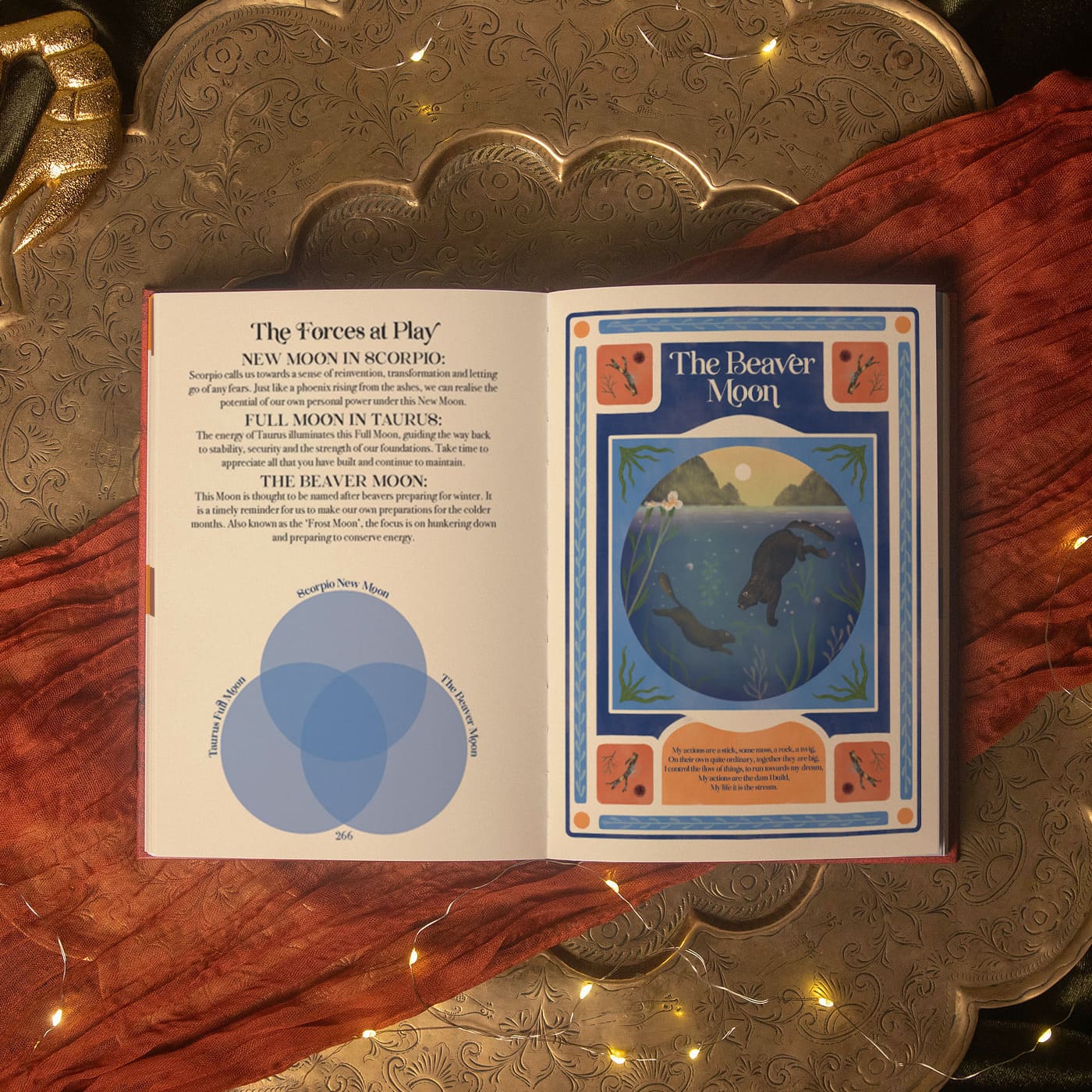 An open book spread showing printed pages titled 'The Forces at Play' and 'The Beaver Moon,' placed on a warm rust-red fabric over an ornate metallic tray with string lights.
