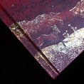 Load image into Gallery viewer, A close-up of a textured dark red or burgundy surface with metallic gold foil detailing, featuring an embossed deer and small floral motif against a black background. Moon Journal 2027
