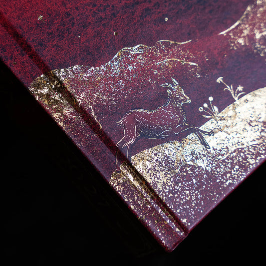 A close-up of a textured dark red or burgundy surface with metallic gold foil detailing, featuring an embossed deer and small floral motif against a black background. Moon Journal 2027