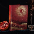 Load image into Gallery viewer, A decorative hardcover 13 moon journal 2027 with a deep red and gold celestial cover design featuring a crescent moon and swirling star pattern, displayed on a dark velvet surface next to a sliced pomegranate and a smoking incense burner.
