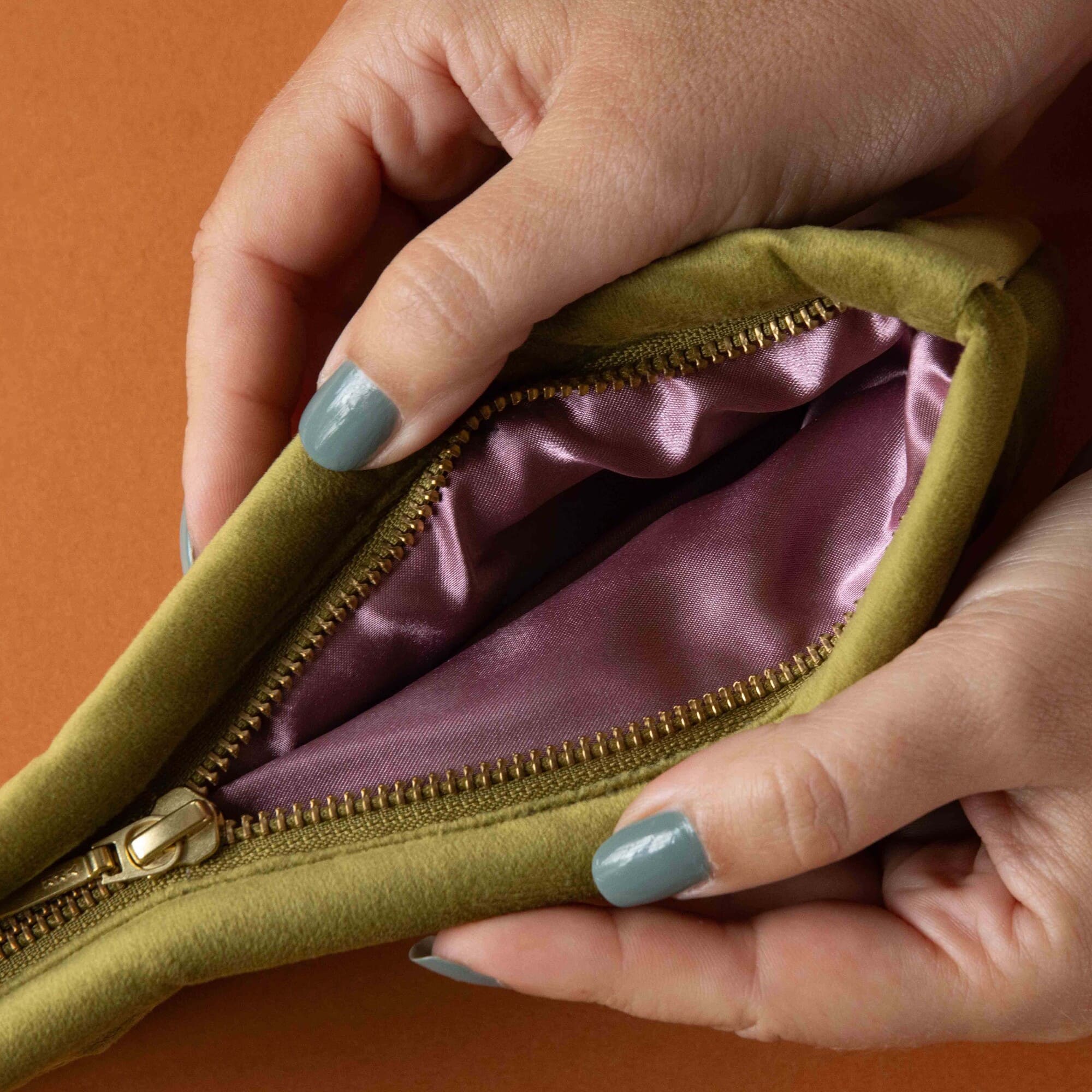 Close-up of hands opening a green zippered pouch with a purple interior on an orange background